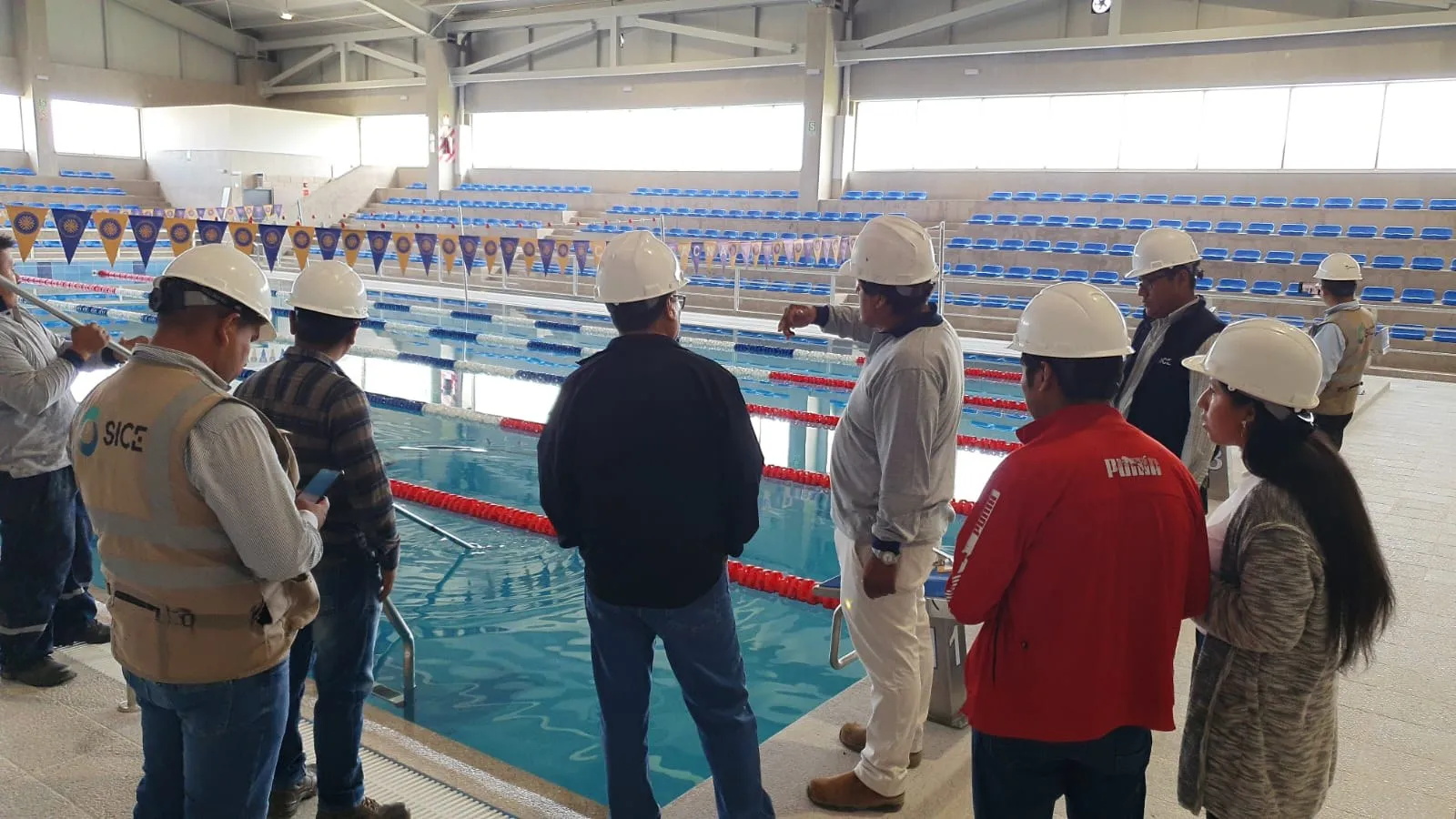 Equipo técnico con cascos revisando una piscina olímpica techada con andariveles y tribunas