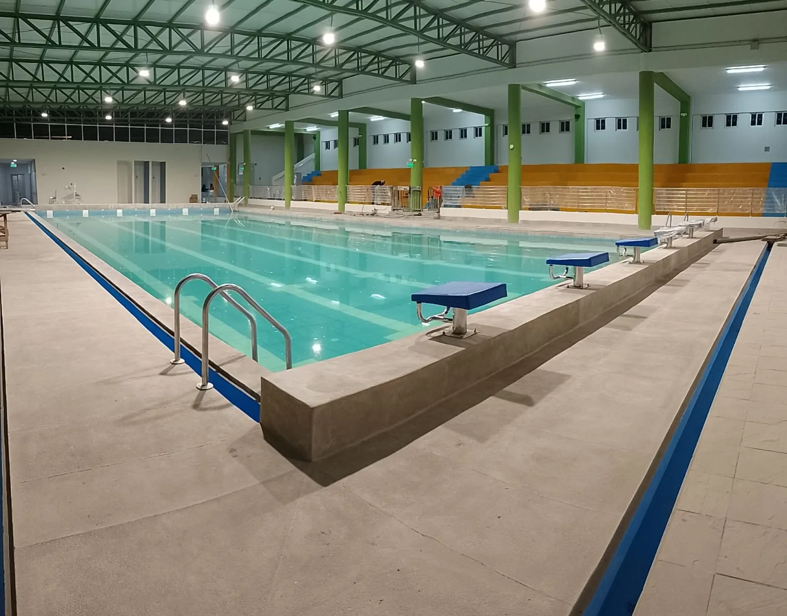 Piscina semi olímpica techada terminada, con pódiums de salida azules, escalera de acero inoxidable y tribunas al fondo
