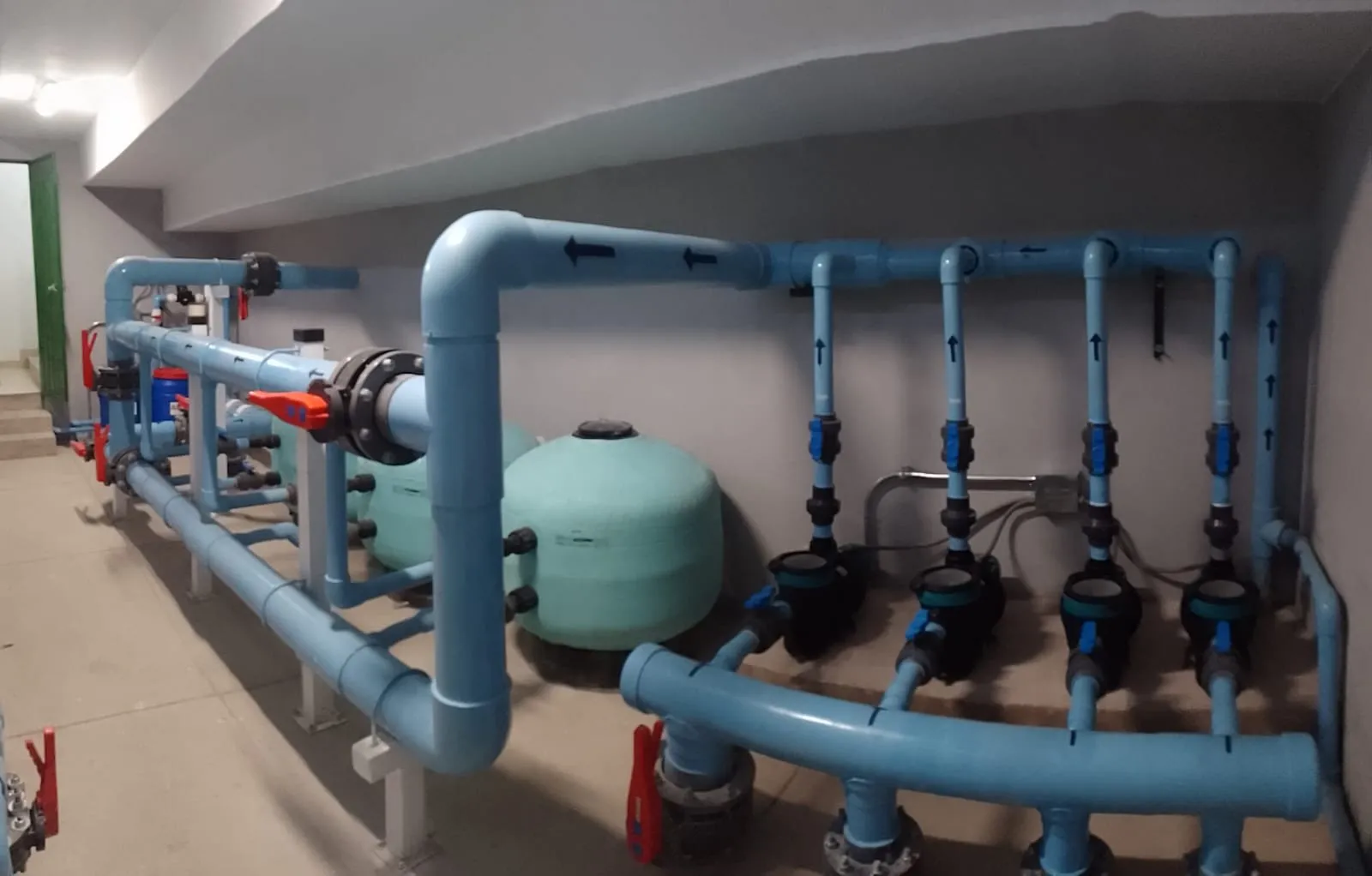 Sala de máquinas de piscina con filtros de arena, cuatro bombas y colectores de PVC señalizados con flechas de flujo